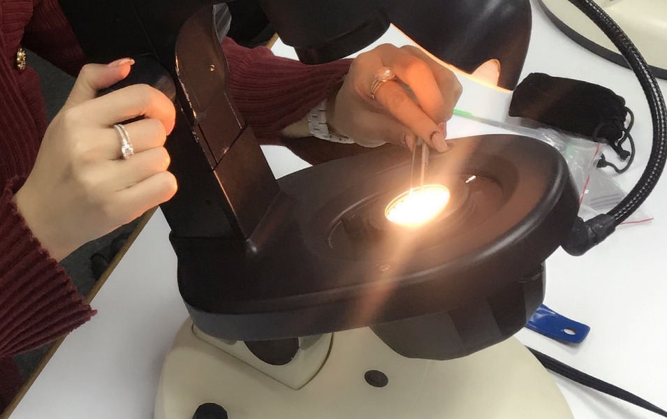 Hands holding a diamond in tweezers under a gem microscope's glowing light