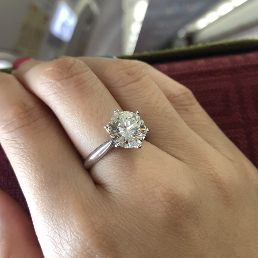 A six-prong round diamond solitaire worn on a hand in an airplane cabin